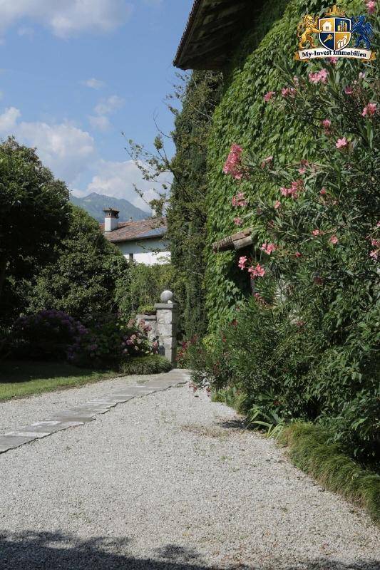 Kiesweg entlang der von Efeu bewachsenen Hausfassade, umgeben von üppiger Vegetation und Bergen im Hintergrund.