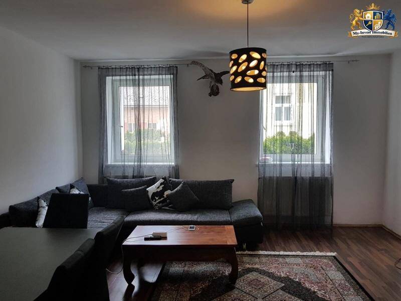 Gemütliches Wohnzimmer mit dunklem Sofa, Holzboden und zwei Fenstern mit transparenten Vorhängen.