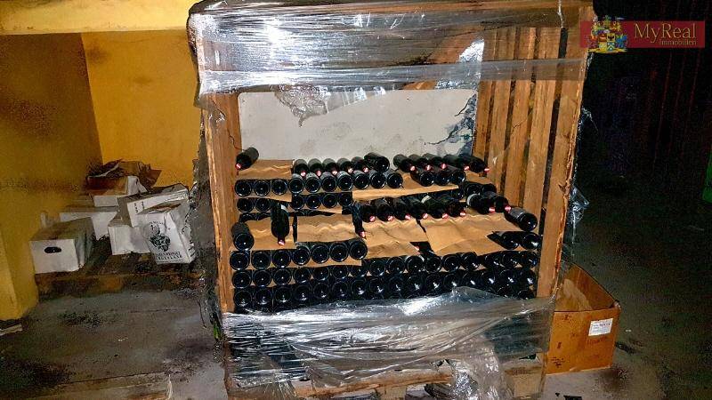 Weinflaschen in einer Holzpalette, mit Folie umwickelt, in einem Lagerraum mit gelblichen Wänden.