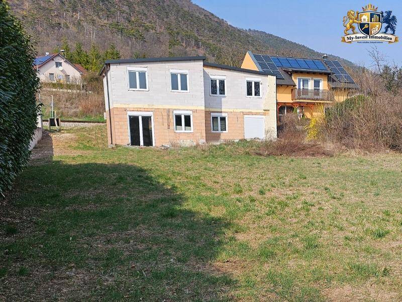 Haus im Rohbau mit unfertiger Fassade und umliegendem Grundstück vor Bergkulisse.