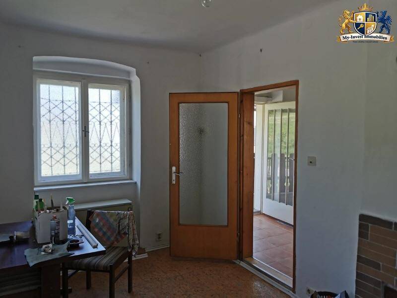 Heller Raum mit Fenster, Holztüren und Fliesenboden, der in einen weiteren Bereich führt.