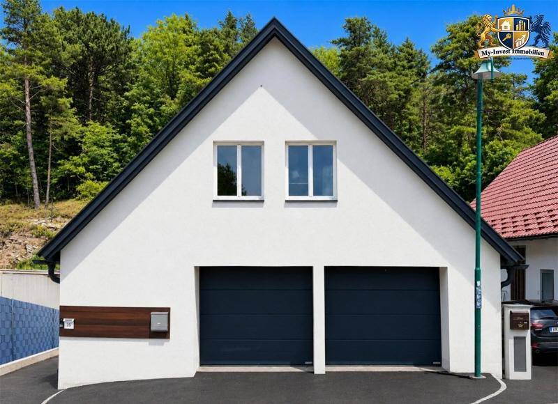 Giebelseite eines modernen Hauses mit Doppelgarage und dunklem Dach, umgeben von Bäumen.