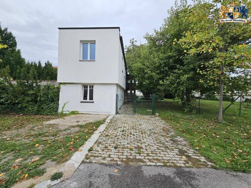 Ansicht des weißen Hauses von vorne mit gepflasterter Auffahrt und einem grünen Zaun.