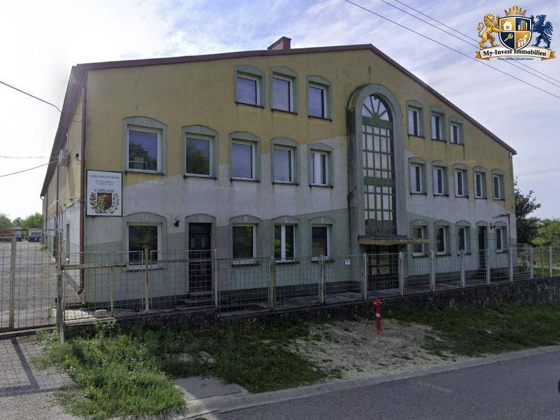 Vorderansicht des Gebäudes mit einem großen Bogenfenster und einem Schild an der Fassade, umgeben von einem Zaun.