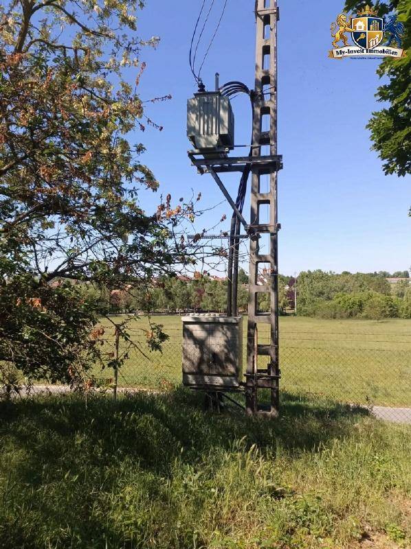 Ein Strommast mit Transformatoren steht auf einem Feld neben einem Zaun unter blauem Himmel.