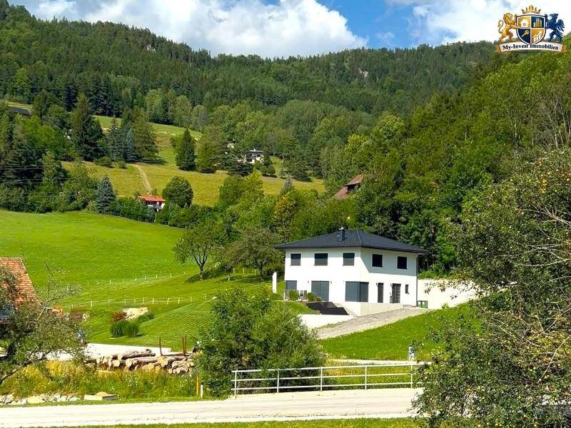 Modernes weißes Haus mit dunklem Dach, eingebettet in eine idyllische Landschaft aus grünen Hügeln und Wäldern.