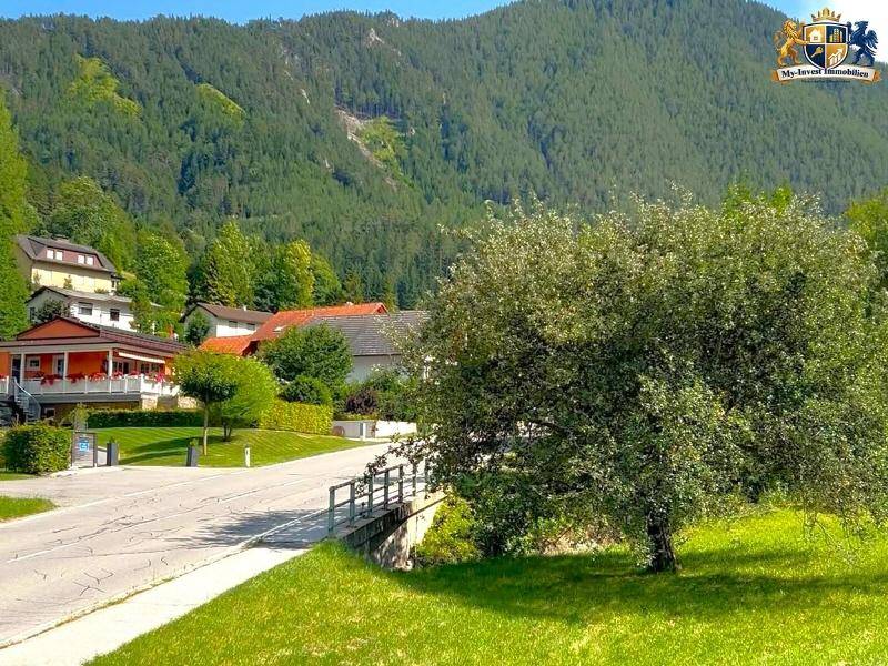 Eine Landstraße schlängelt sich durch eine Siedlung mit Häusern, umgeben von grünen Wiesen und Bergen.