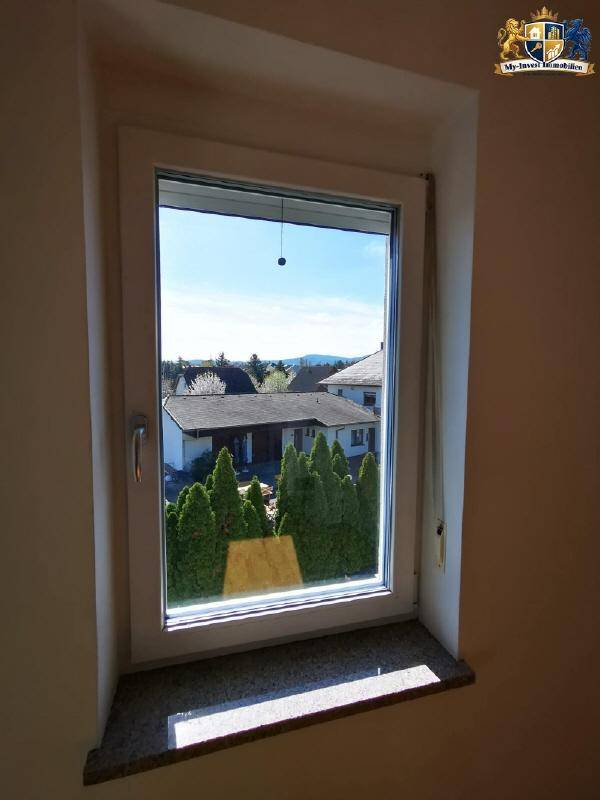 Fensterblick auf grüne Bäume und die Dächer der umliegenden Häuser in der Ferne.