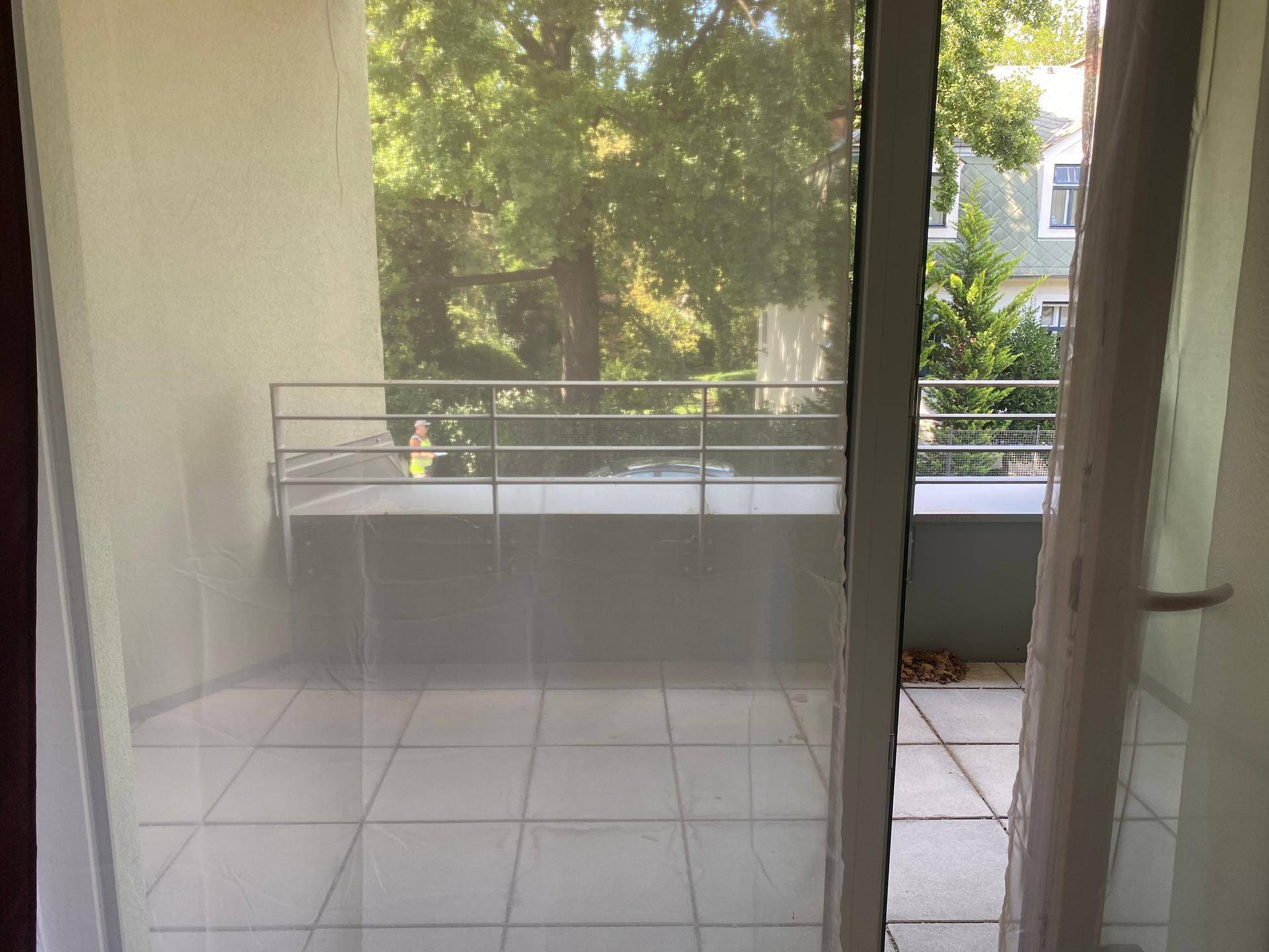 Blick vom Innenraum auf einen Balkon mit Fliesenboden und Metallgeländer, umgeben von Bäumen.