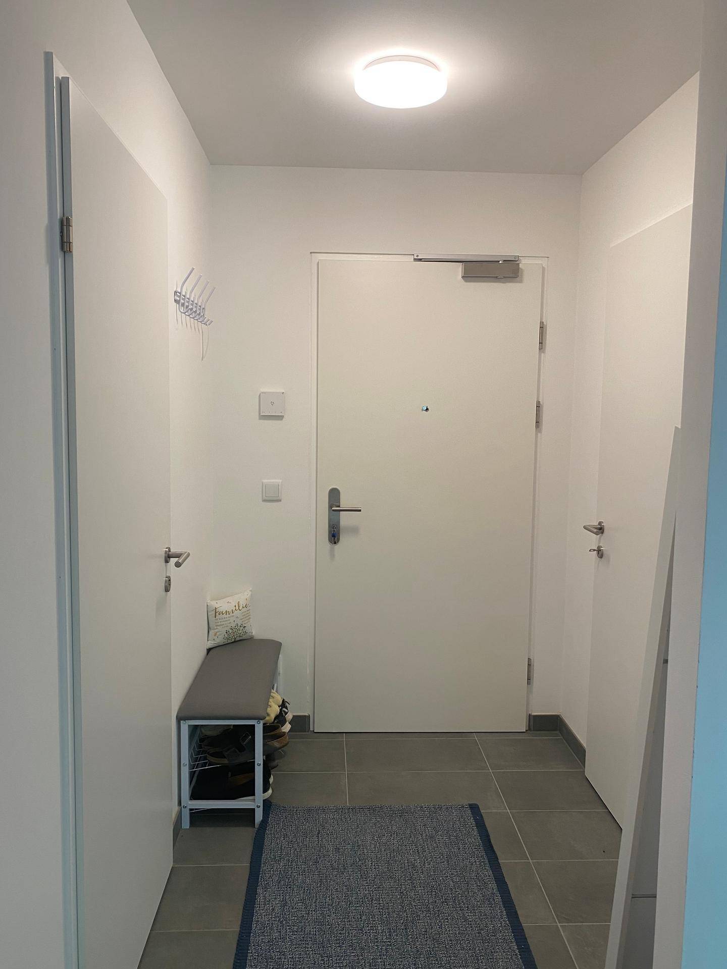 Heller Eingangsbereich mit weißer Tür, grauen Fliesen und kleiner Sitzbank.