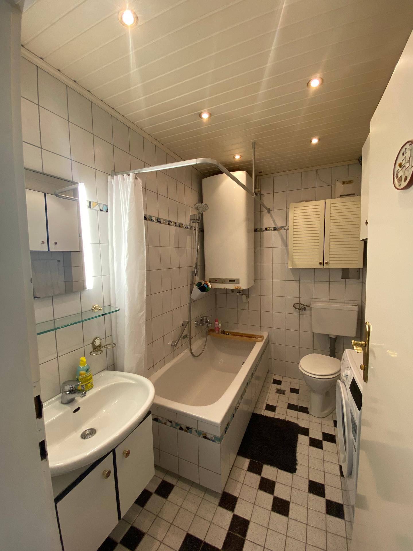 Badezimmer mit Badewanne, Duschvorhang, WC und einem schwarz-weiß gefliesten Boden im 80er-Jahre-Stil.