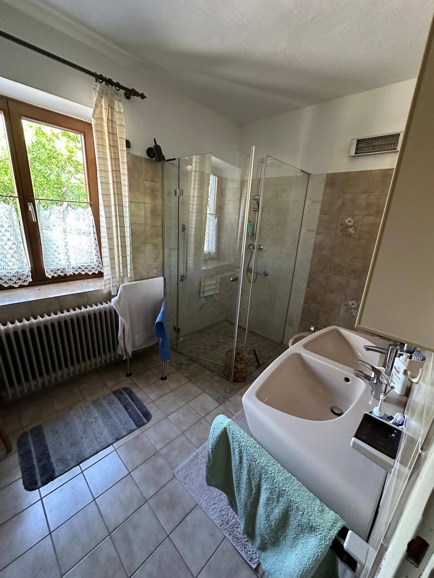 Badezimmer mit Fliesenboden, Doppelwaschbecken und großer Duschkabine, hell durch Fenster.