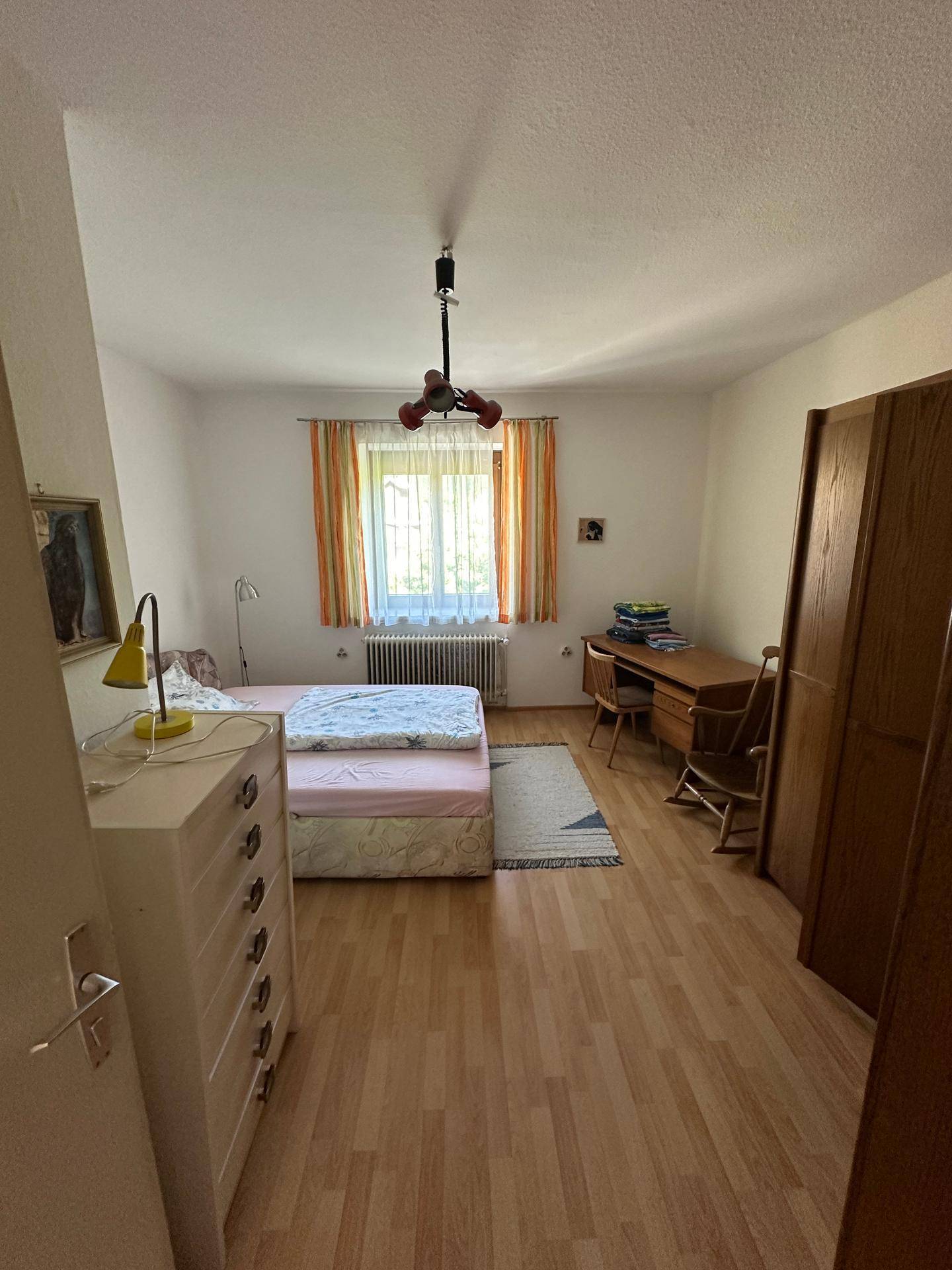 Einfaches Schlafzimmer mit Holzoptikboden, Schreibtisch und Kleiderschrank, hell und funktional.