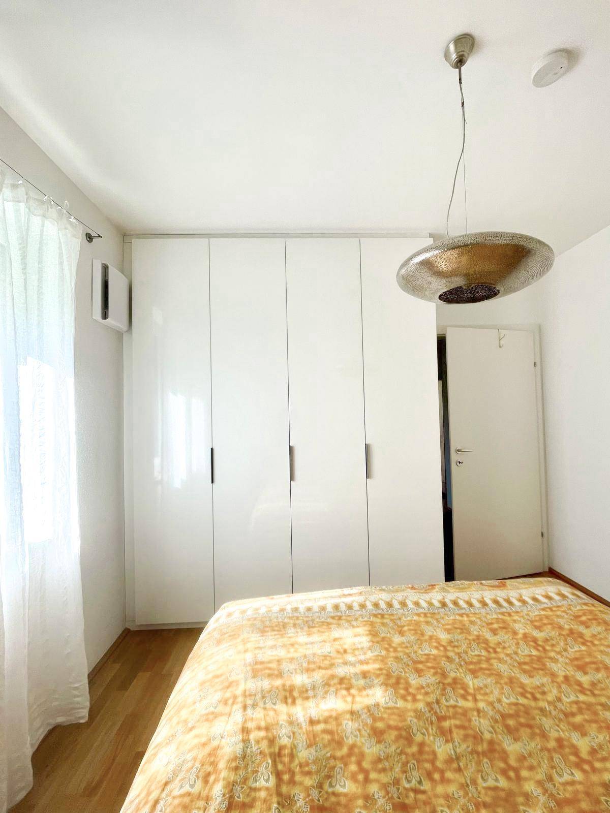 Helles Schlafzimmer mit weißem Einbauschrank, Doppelbett und Fenster mit Vorhängen.