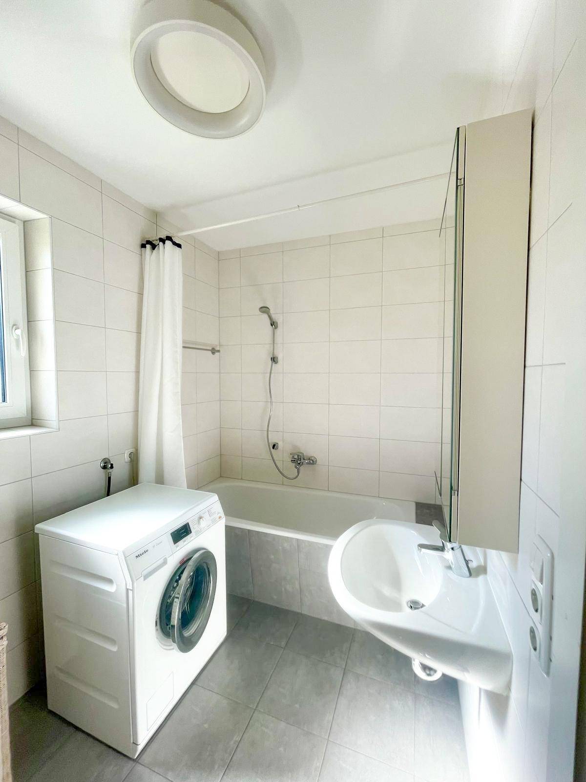 Helles Badezimmer mit Badewanne, Waschmaschine, Waschbecken und Fenster.