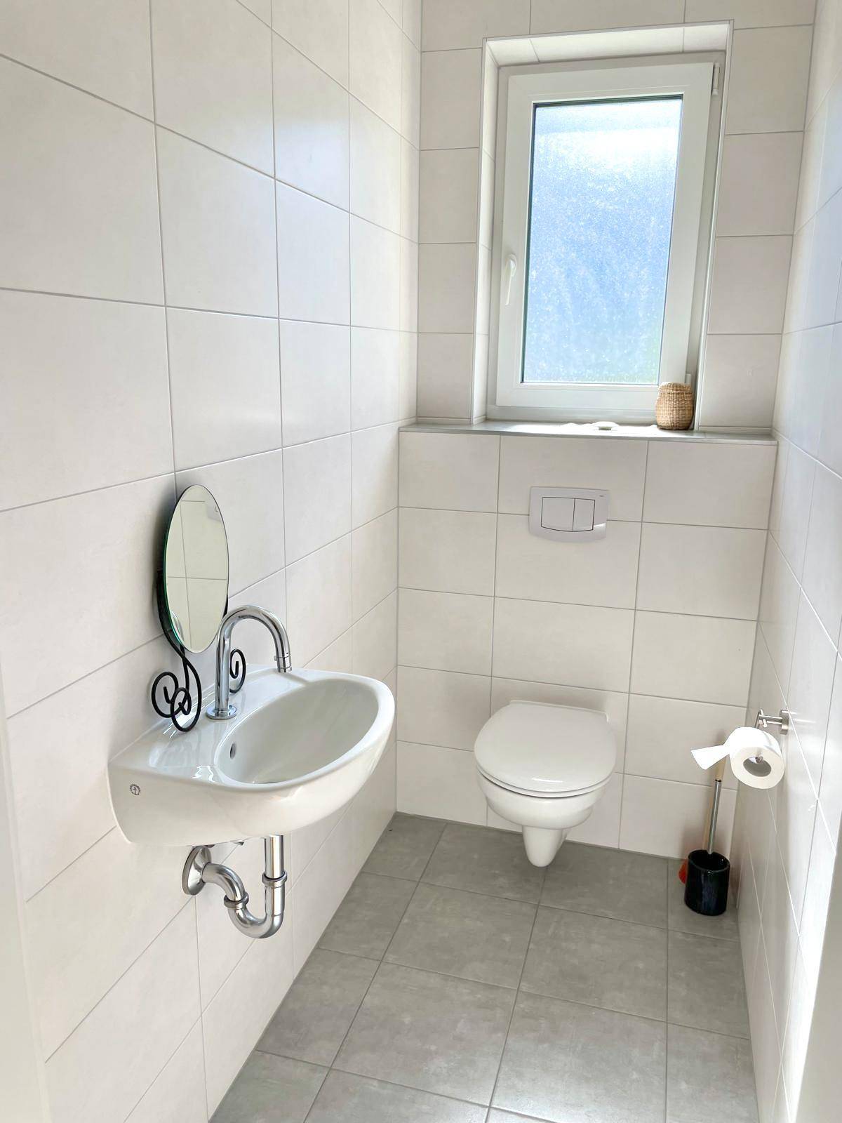 Modernes Gäste-WC mit wandhängender Toilette, kleinem Waschbecken und Fenster für Belüftung.