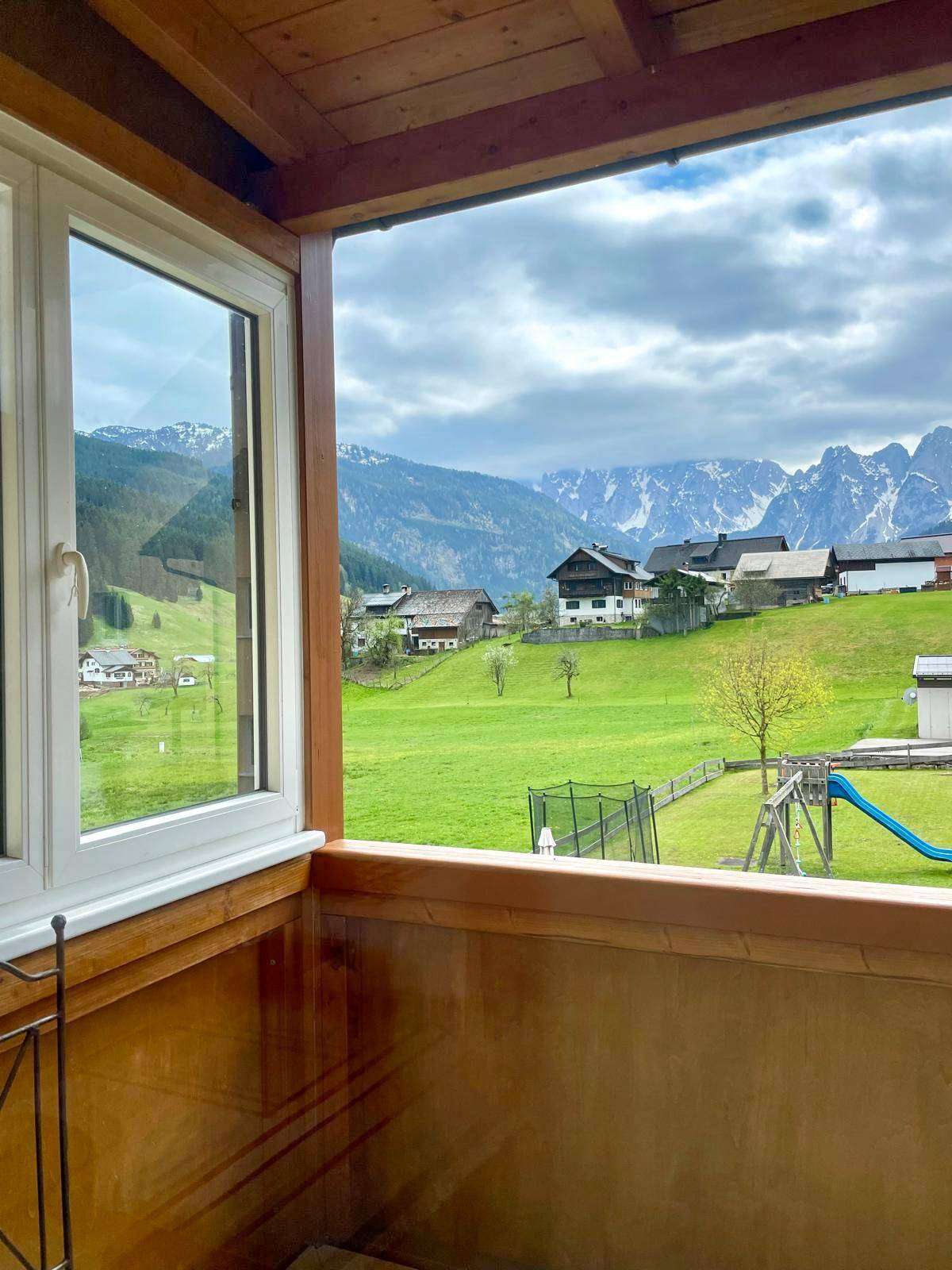 Panoramablick von einem Holzbalkon auf grüne Hügel, Berge und einen Kinderspielplatz.