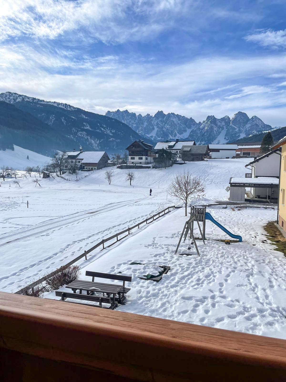 Winterlicher Ausblick von einem Balkon auf schneebedeckte Berge, Häuser und einen Spielplatz.