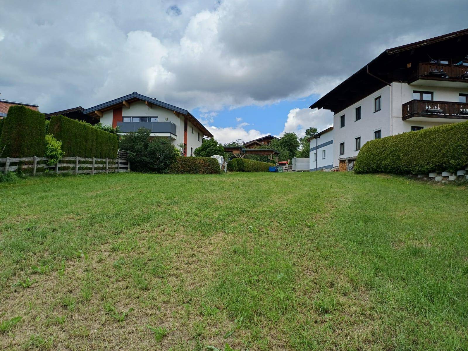 Grüne Rasenfläche mit Hecken und zwei Wohnhäusern im Hintergrund unter einem bewölkten Himmel.