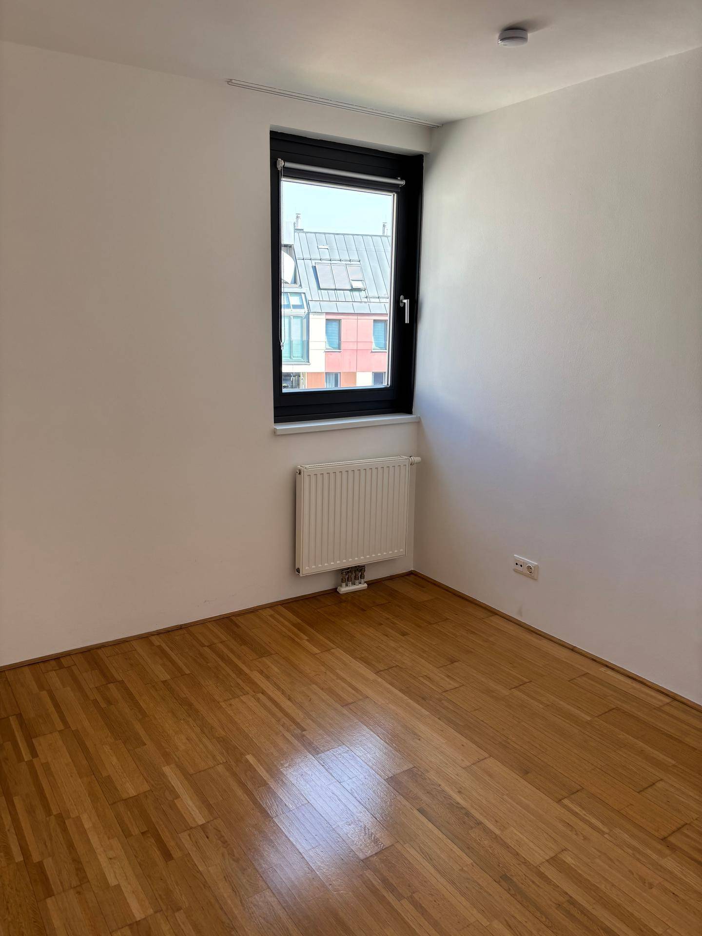 Leeres Schlafzimmer mit Parkettboden und einem Fenster, das auf die umliegenden Gebäude blickt.
