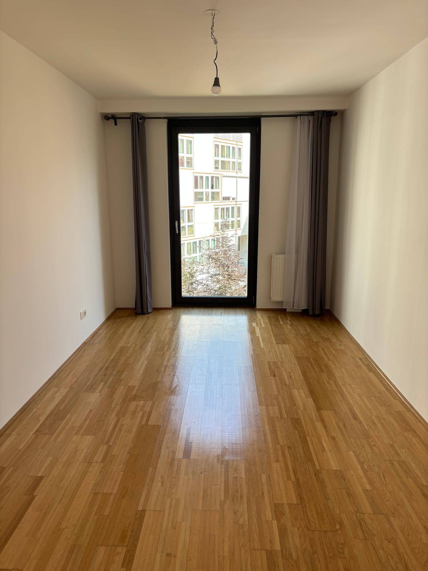 Leeres Schlafzimmer mit Parkettboden und großem Fenster mit Vorhängen.