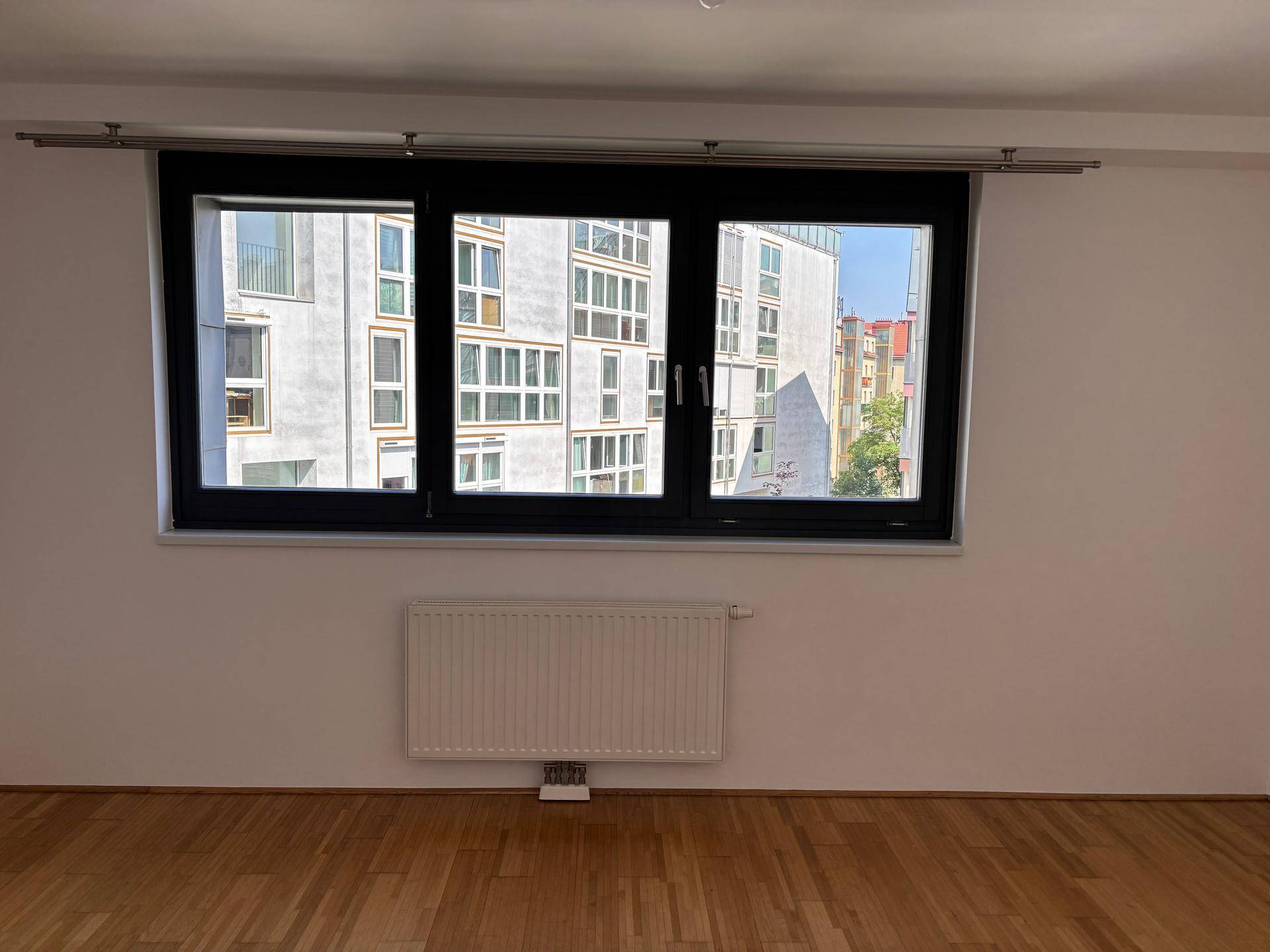 Helles Schlafzimmer mit Parkettboden und breitem Fenster mit Blick auf die Nachbarschaft.