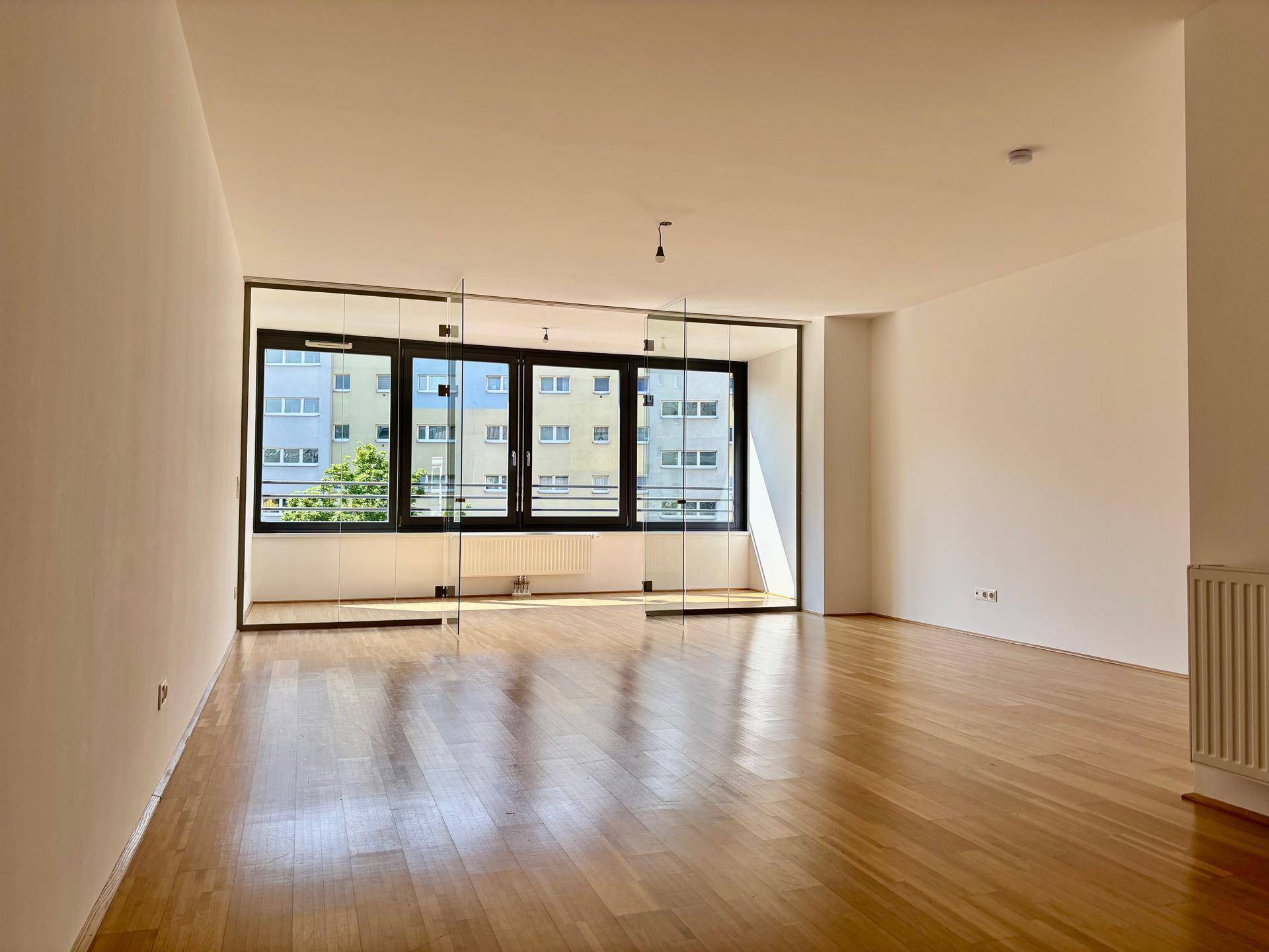 Helles Wohnzimmer mit Parkettboden, großen Fenstern und Blick auf die umliegenden Gebäude.