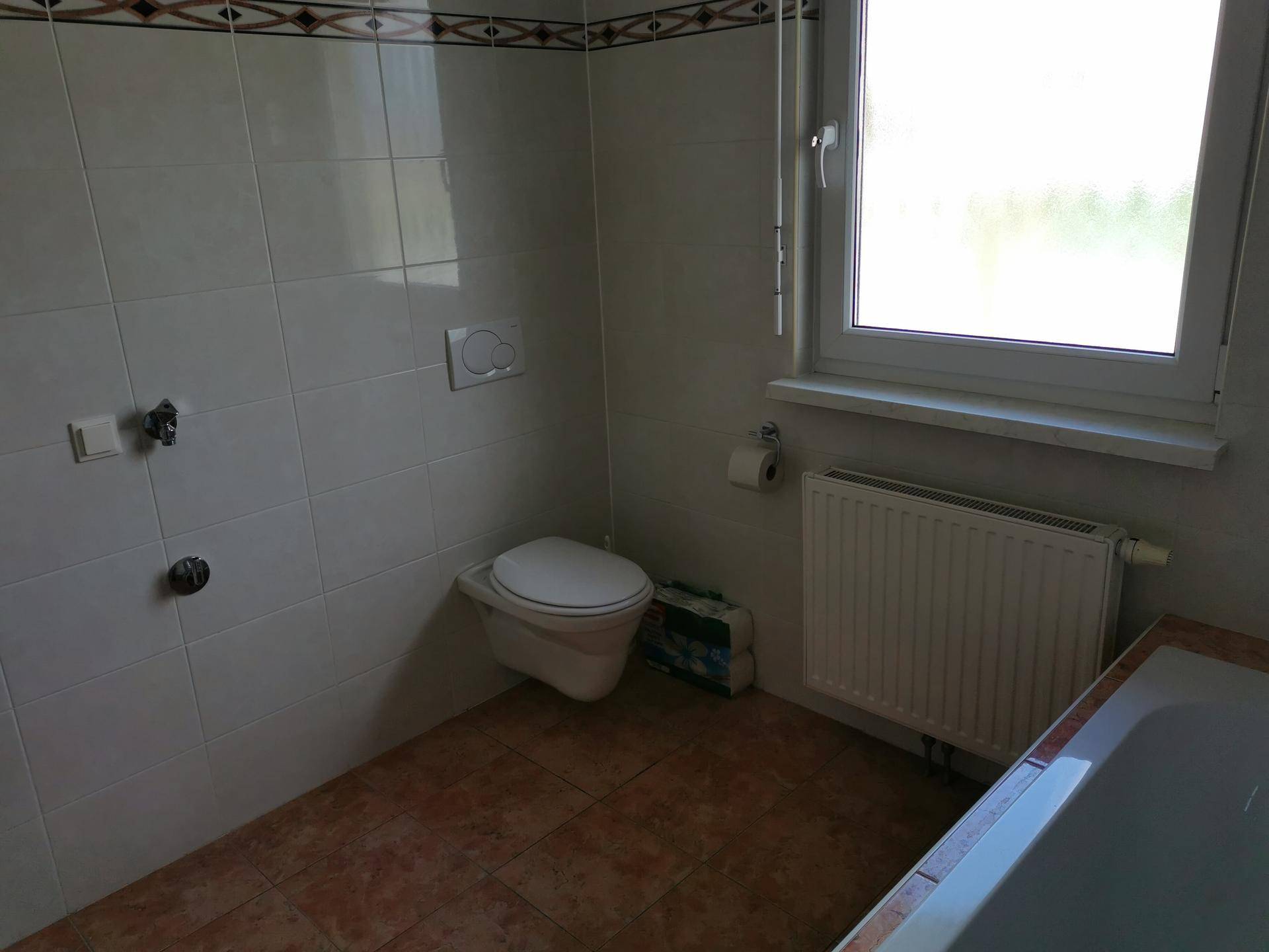 Badezimmer mit Badewanne, Wand-WC, Fenster und terracottafarbenen Fliesenboden.