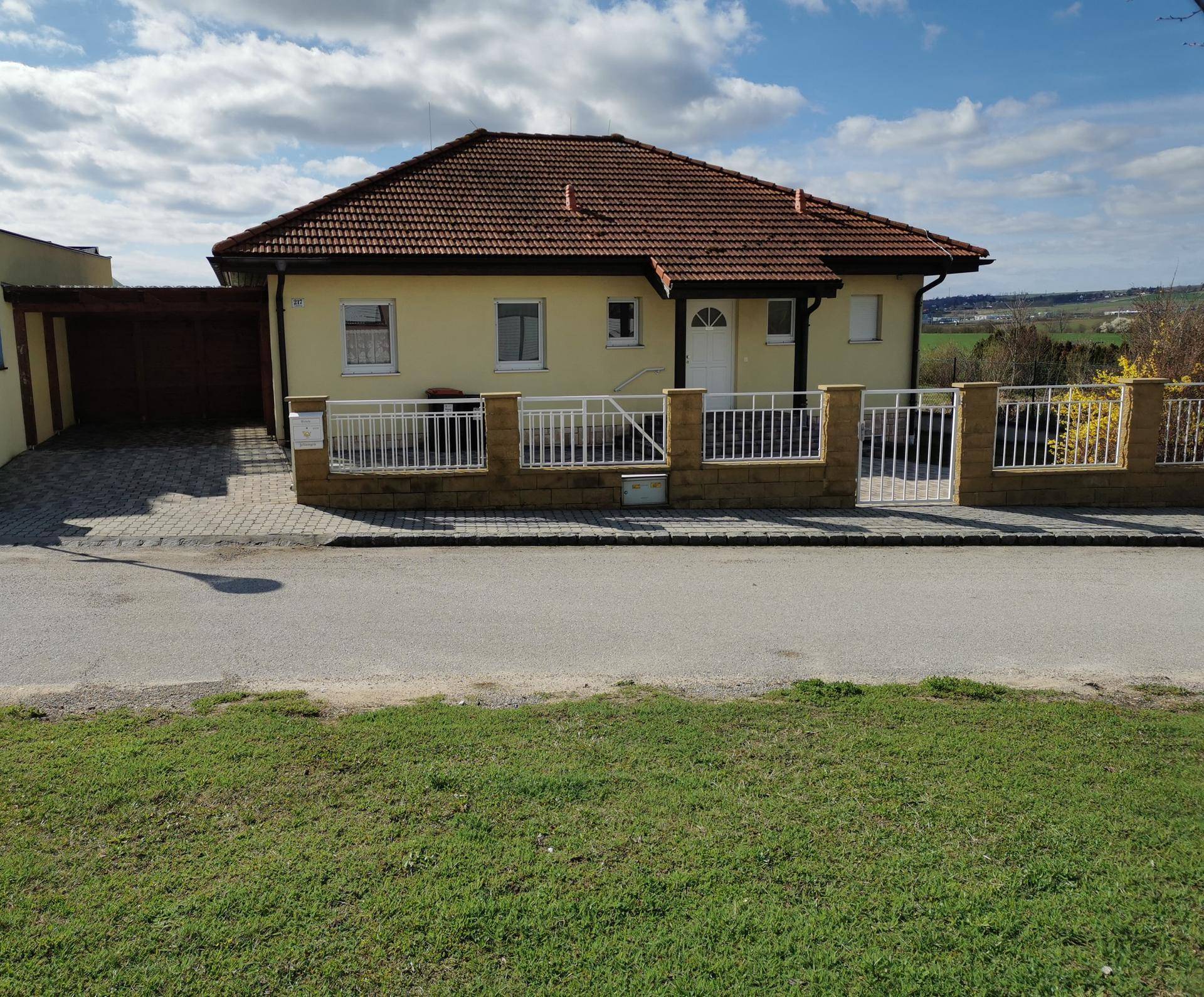 Vorderansicht des Einfamilienhauses mit gepflasterter Einfahrt, Zaun und Satteldach unter blauem Himmel.