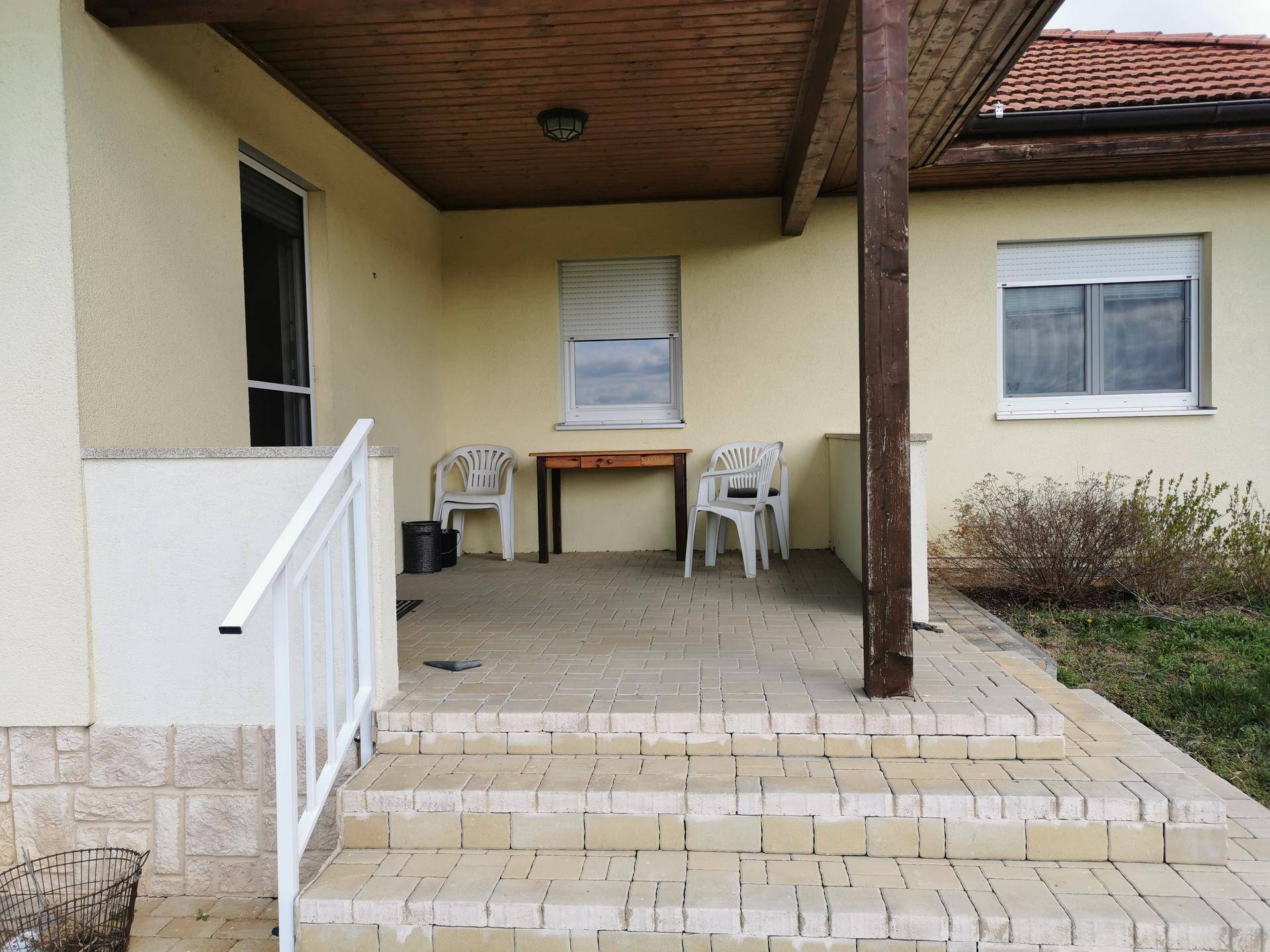 Überdachte Terrasse mit Fliesenboden und einfacher Sitzgelegenheit vor dem Haus.
