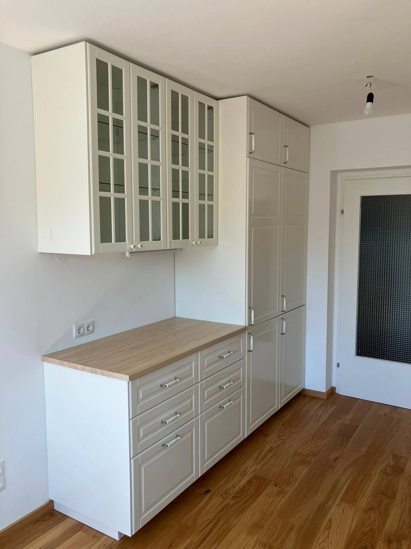 Erweiterte Küchenzeile mit weißen Schränken, Glasfronten und Platz für einen Kühlschrank.