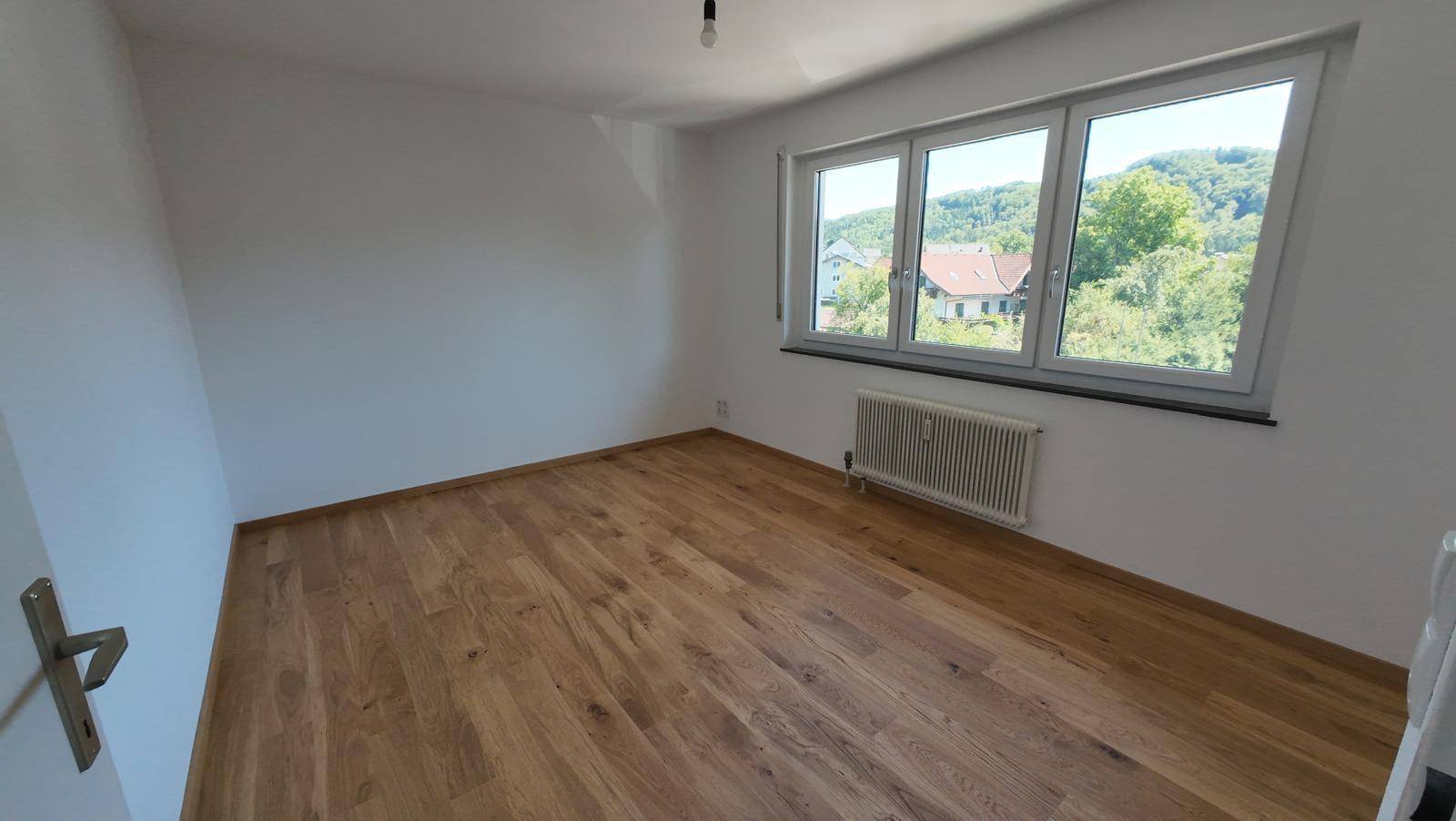 Helles Schlafzimmer mit Parkettboden und großem Fenster mit Blick auf die Umgebung.