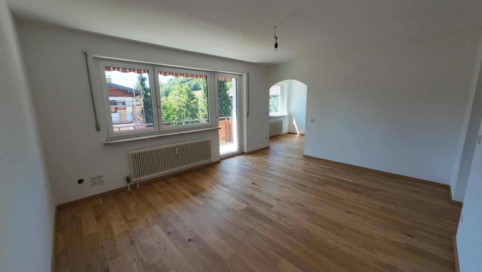 Geräumiges Wohnzimmer mit hellem Parkettboden, großem Fenster und Zugang zum Balkon mit Grünblick.