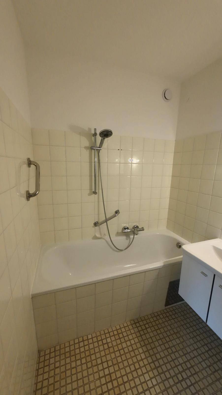 Badewanne mit Duschfunktion und Haltegriff in einem gefliesten Badezimmer.