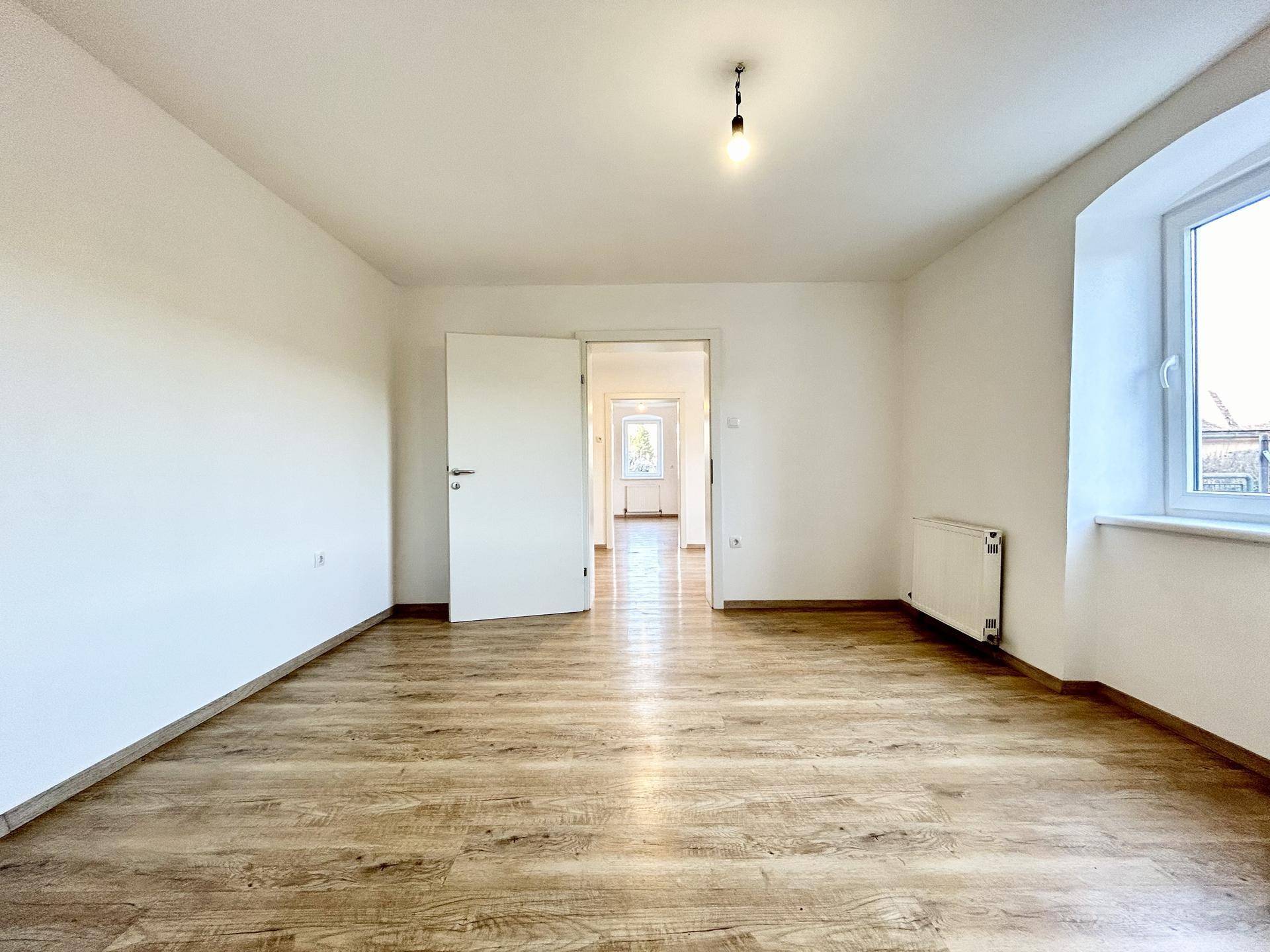 Geräumiger, leerer Raum mit Laminatboden und Blick durch eine offene Tür in einen weiteren Bereich.
