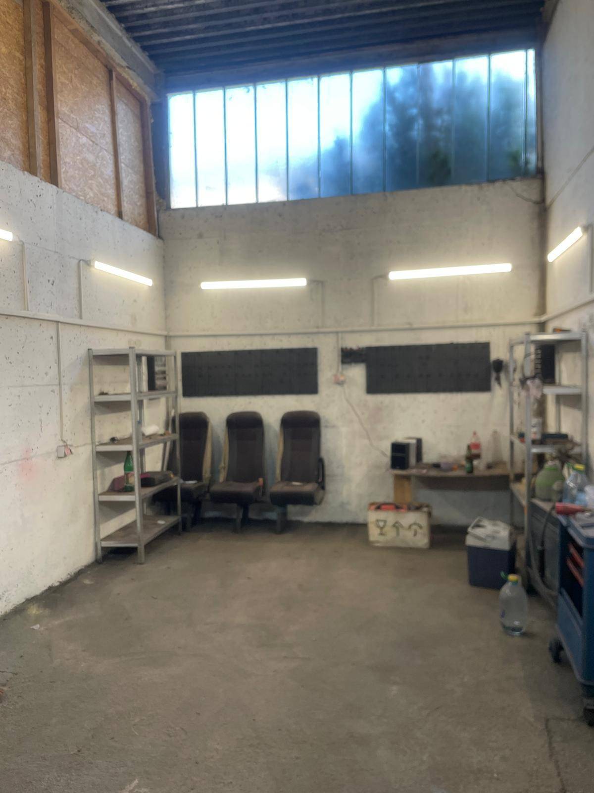 Geräumiger Werkstattraum mit einfacher Einrichtung, Metallregalen und hohen Oberlichtern.