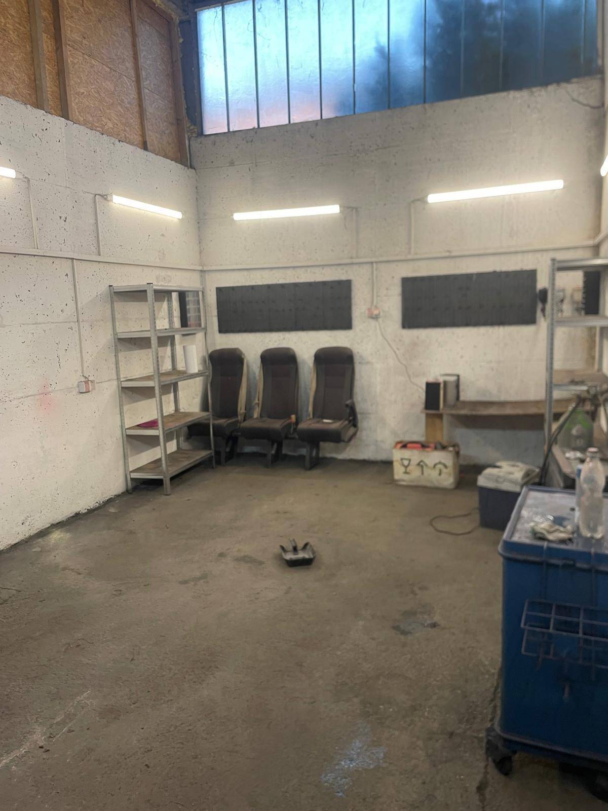 Geräumiger Werkstattraum mit einfacher Einrichtung, Metallregalen und hohen Oberlichtern.