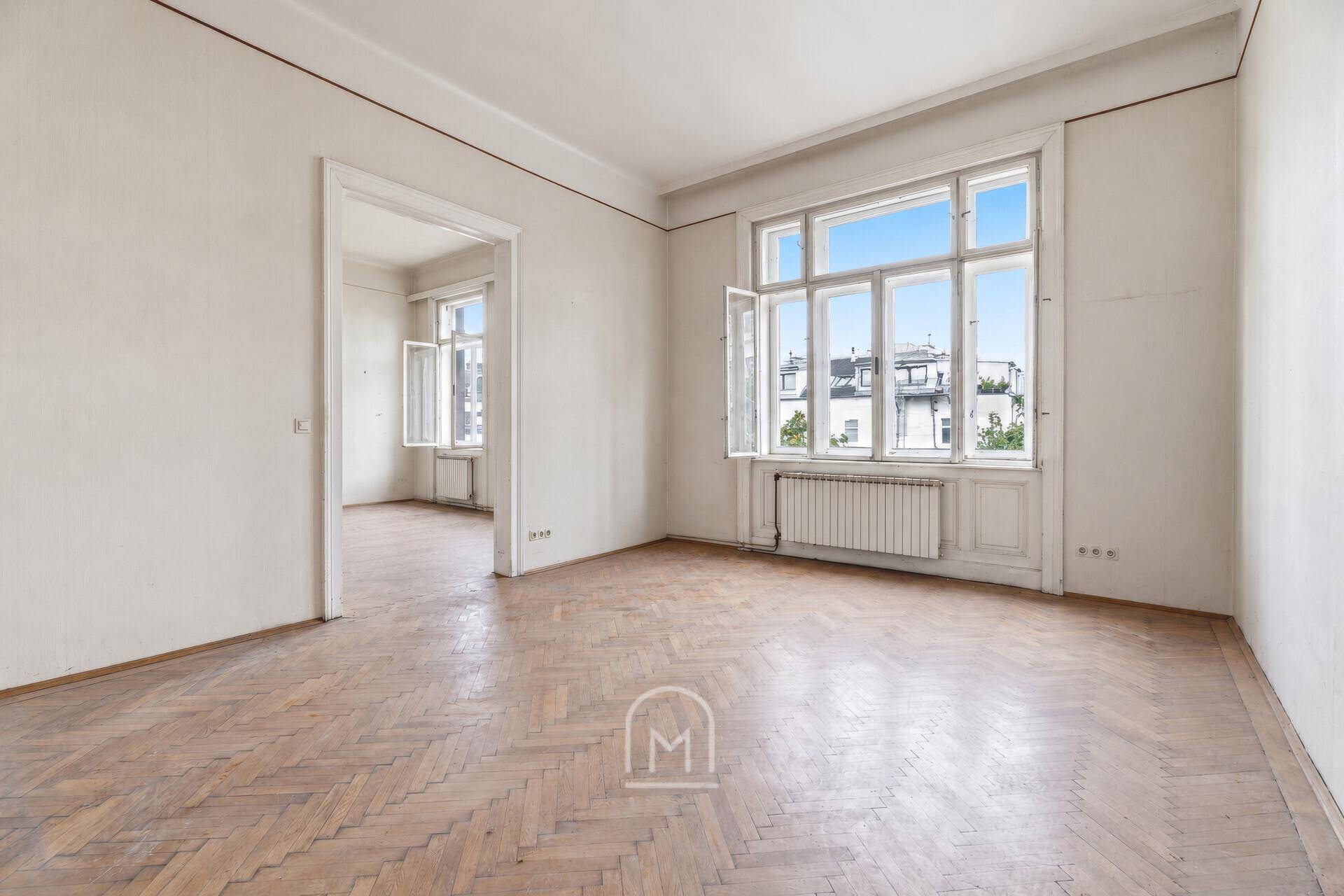 Helles Zimmer mit Fischgrätparkett und großen Fenstern, die viel Tageslicht hereinlassen.