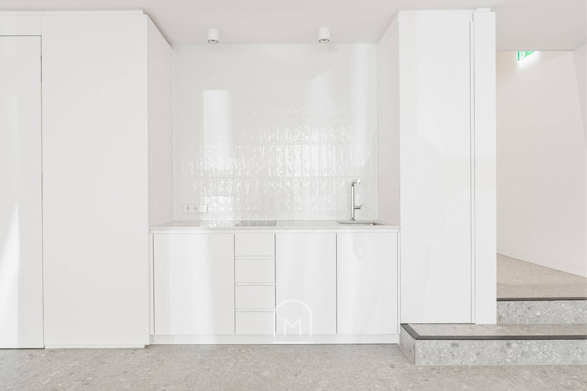 Moderne, weiße Teeküche mit Spüle, Kochfeld und glänzenden Fliesen an der Wand.