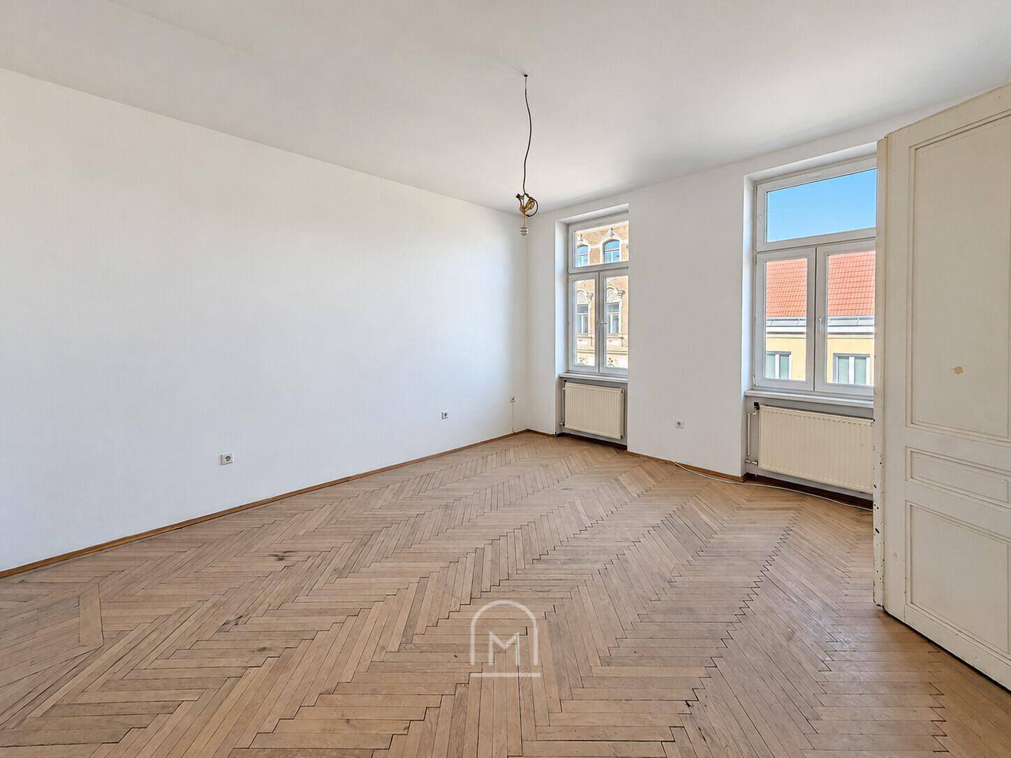 Geräumiges Zimmer mit klassischem Fischgrätparkett und viel Tageslicht durch zwei große Fenster.