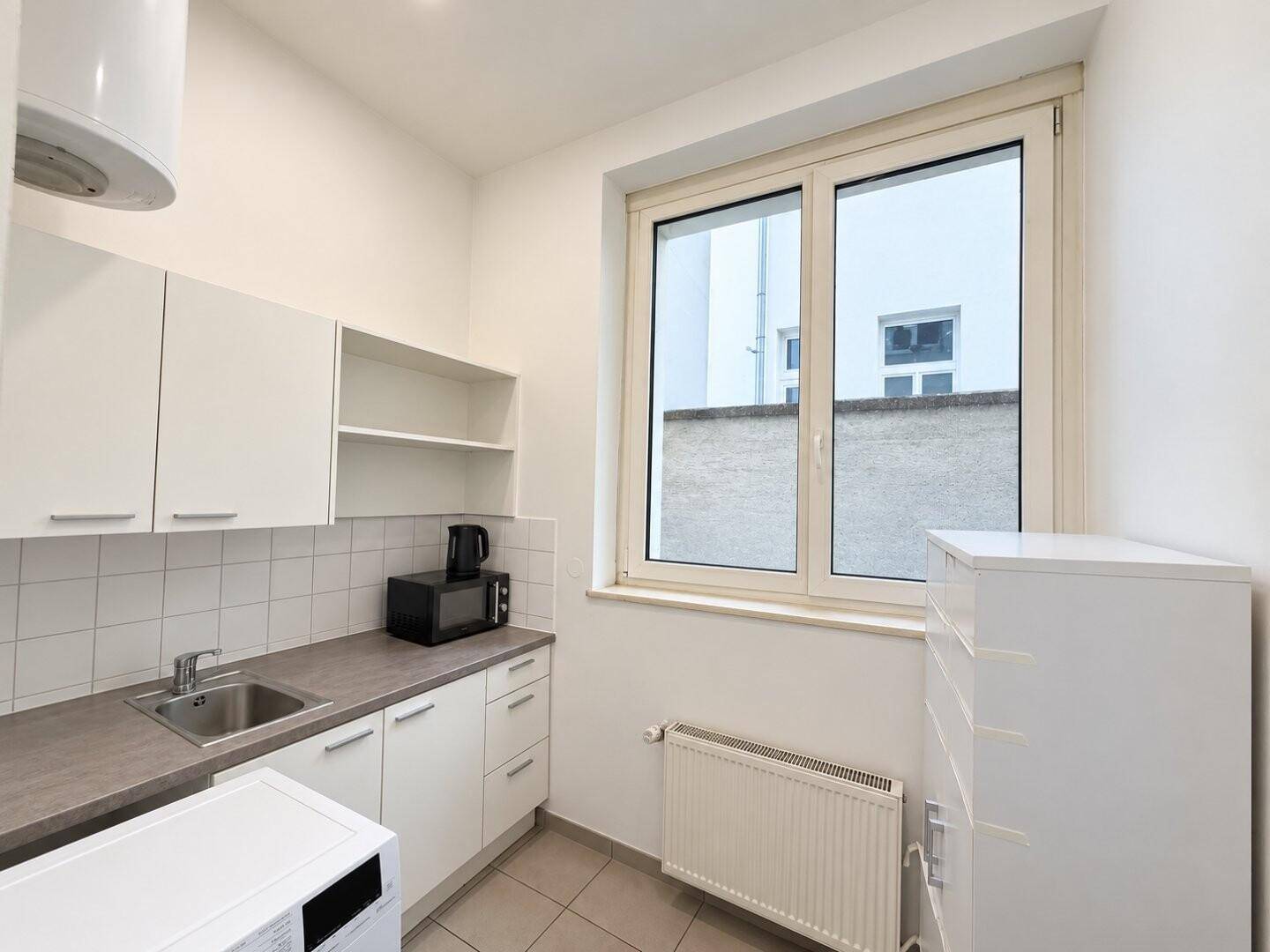 Kleine Küchenzeile mit weißen Schränken, Spüle, Mikrowelle und einem Fenster für Tageslicht.