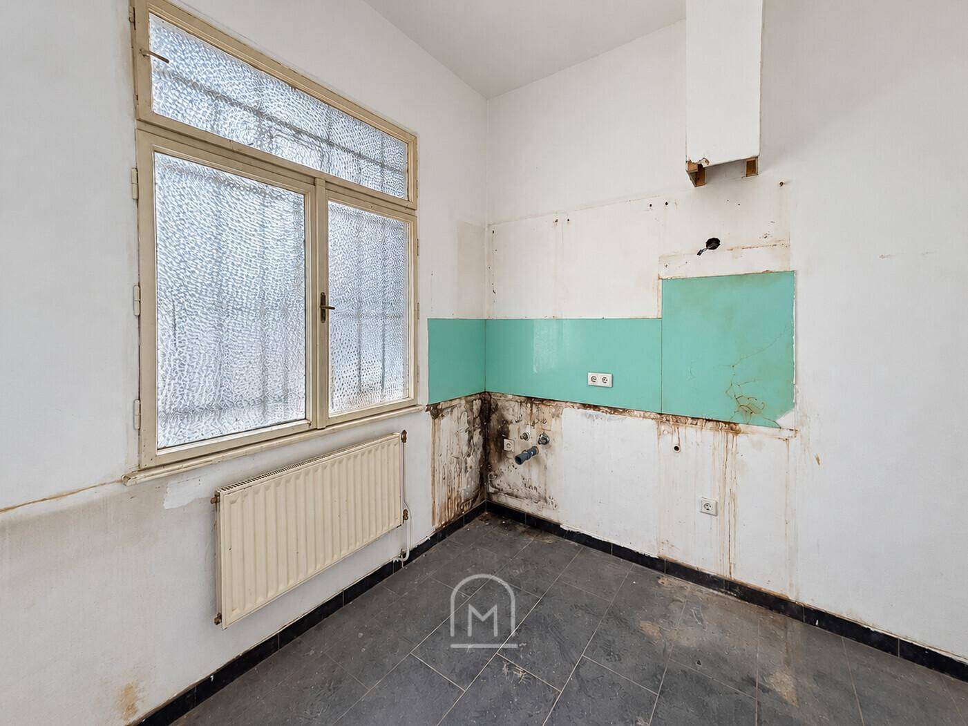 Renovierungsbedürftige Küche mit alten Fliesen, sichtbaren Leitungen und einem Fenster für Tageslicht.