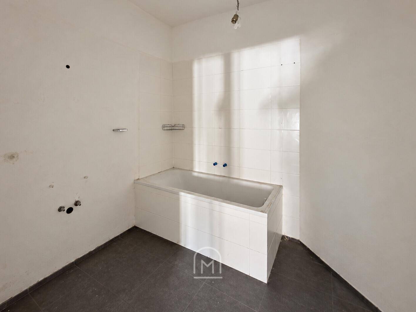 Badezimmer im Rohzustand mit eingebauter Badewanne und dunklen Fliesen, benötigt Renovierung.