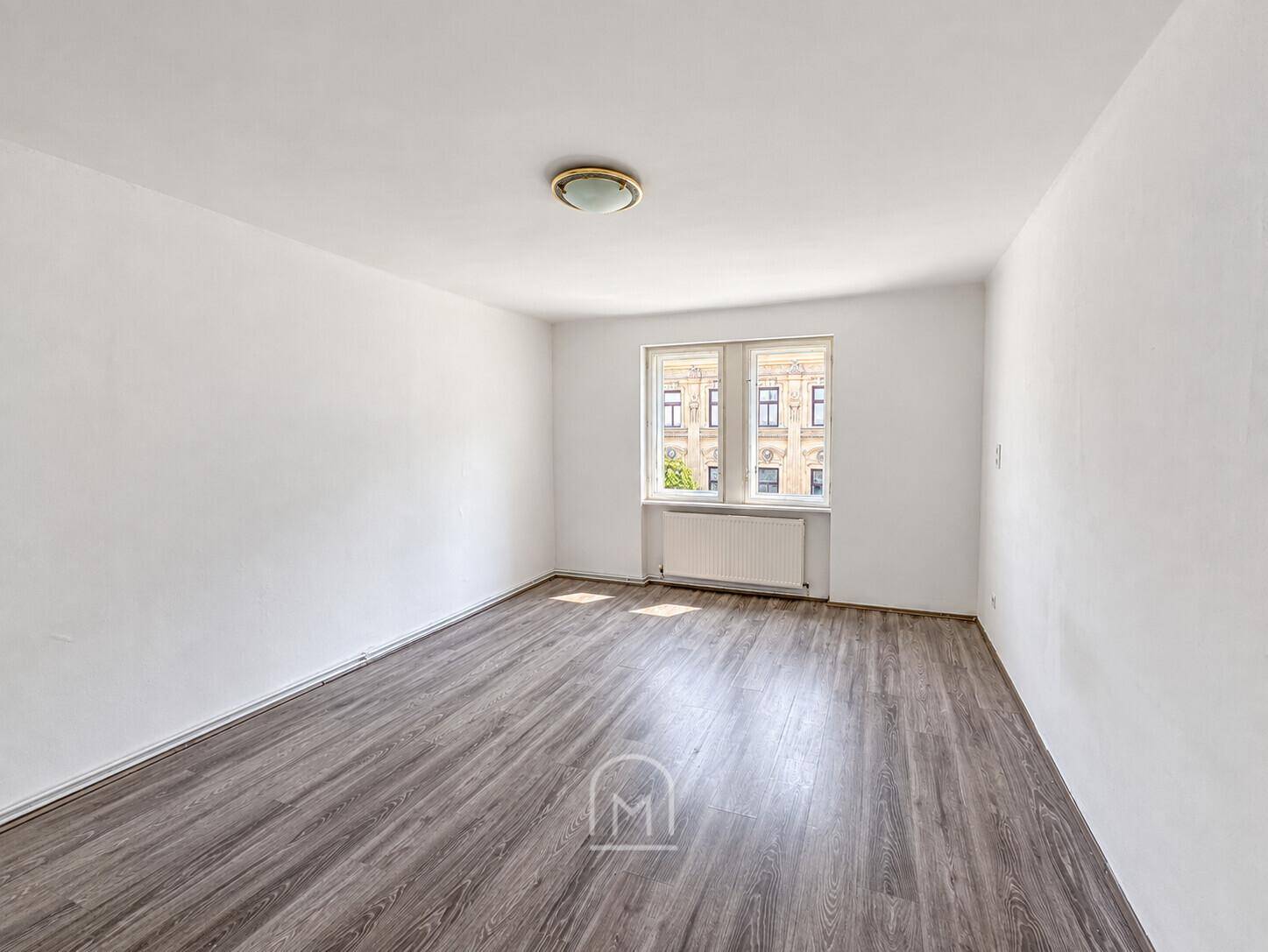 Leeres Wohnzimmer mit Laminatboden und einem großen Fenster, das viel Tageslicht hereinlässt.