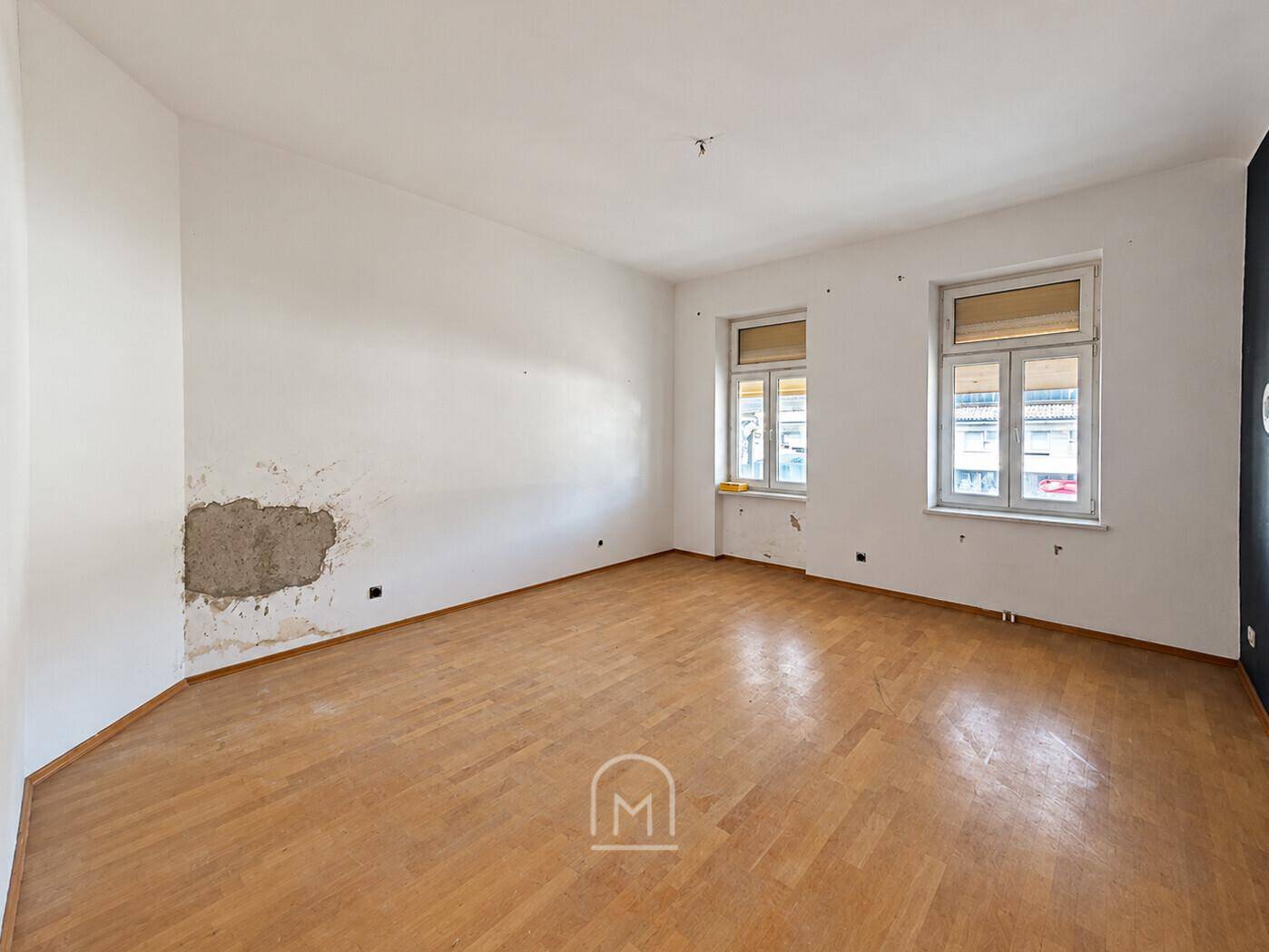 Unmöblierter Raum mit Parkettboden und sichtbaren Putzschäden an einer Wand, renovierungsbedürftig.