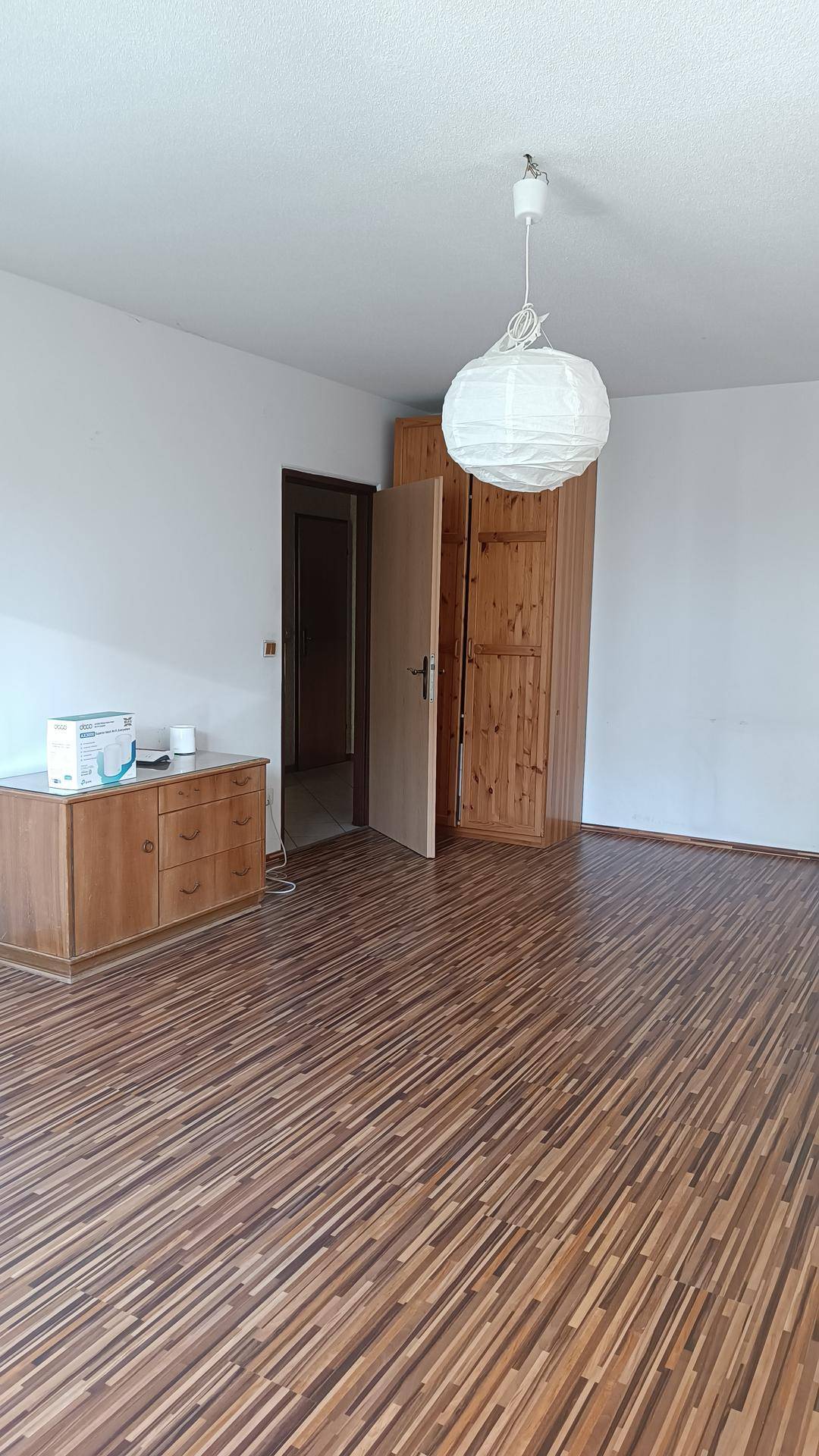 Geräumiger Wohnraum mit Holzstreifenboden, weiß gestrichenen Wänden und einer Holztür, die zu einem weiteren Raum führt.