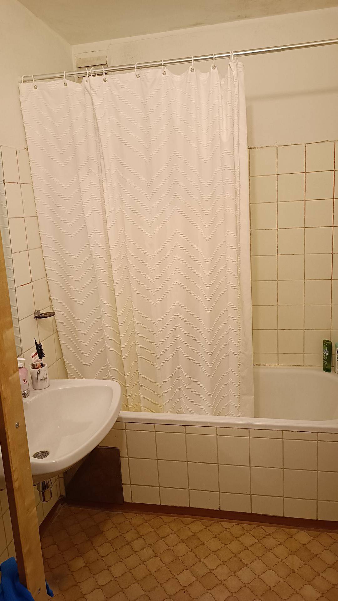 Badezimmer mit Badewanne, Duschvorhang, Waschbecken und hellen, gemusterten Fliesen an Wänden und Boden.