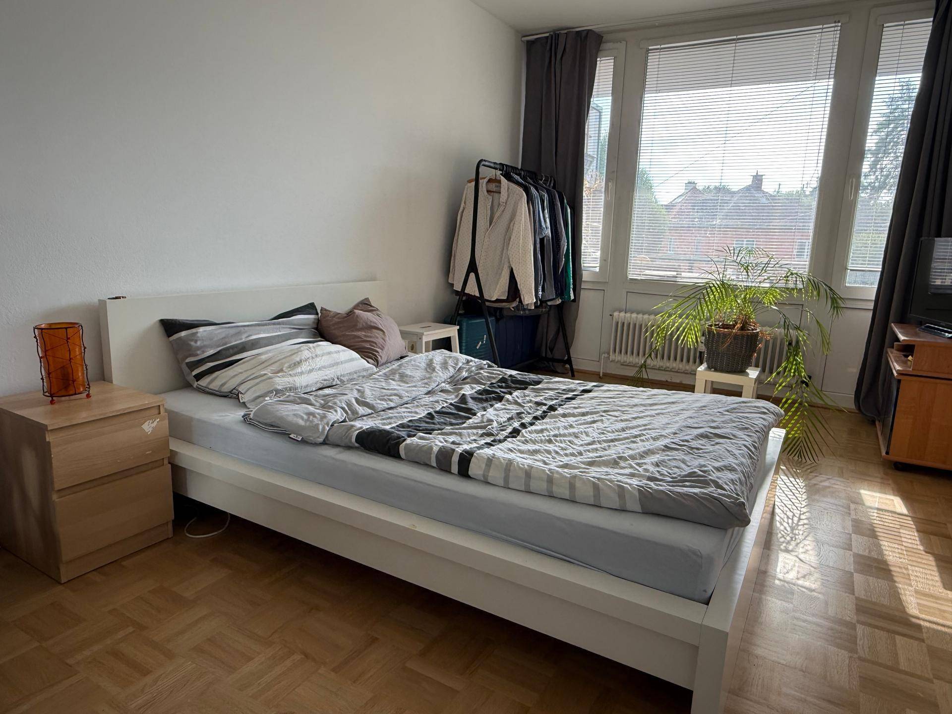Helles Schlafzimmer mit Bett, Nachttisch und Kleiderstange, durchflutet von natürlichem Licht.