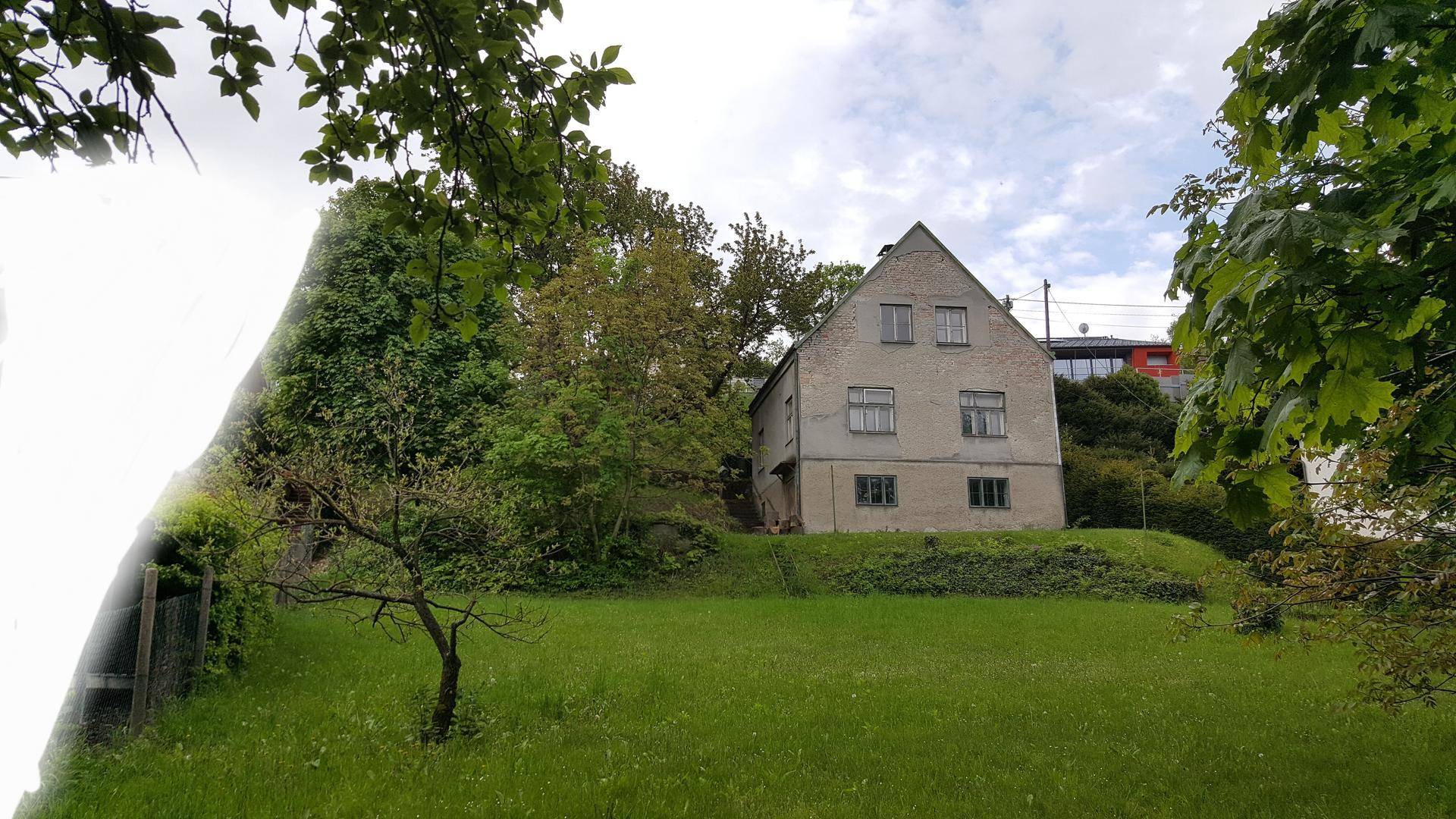 Hausfassade mit umgebendem Grün und Bäumen unter bewölktem Himmel.