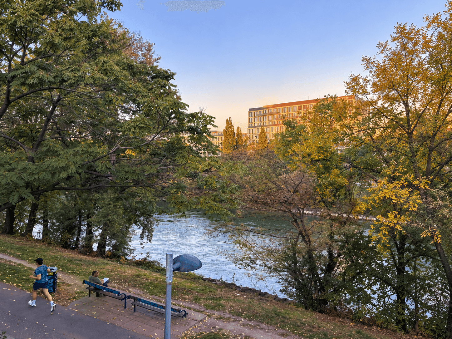 Malerischer Blick auf einen Fluss mit üppiger Vegetation und Gebäuden im Hintergrund bei Sonnenuntergang.